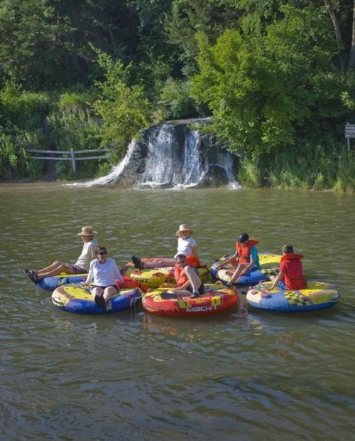 A group of people floating on colorful inner tubes in a river near a small waterfall and lush greenery.