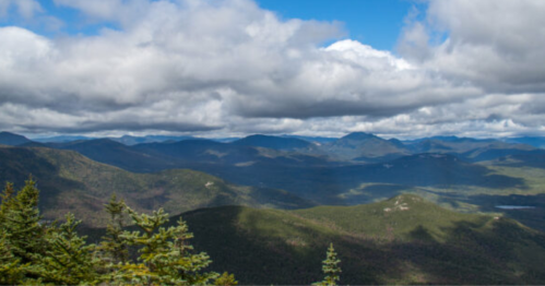 A panoramic view of mountains and valleys under a cloudy blue sky, with lush greenery in the foreground.