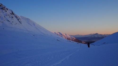 A snowy mountain landscape at sunset, with a lone figure walking along a path in the foreground.
