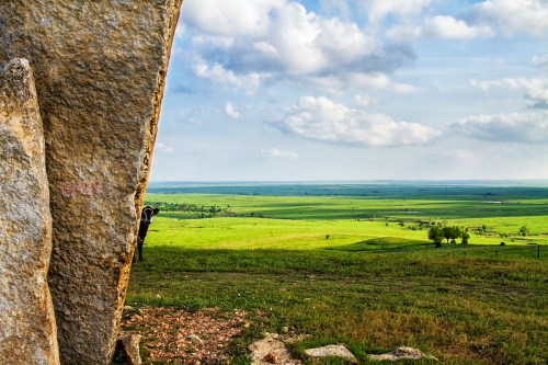 A vast green landscape stretches under a cloudy sky, viewed from behind a large rock formation.