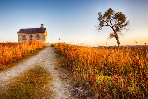 A rustic stone building beside a winding path, surrounded by tall grass and a solitary tree under a clear sky.
