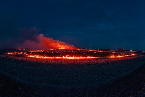 A nighttime landscape of a field ablaze, with flames and smoke rising against a dark sky.