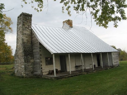A rustic wooden house with a stone chimney, featuring a metal roof and a porch, surrounded by green grass and trees.