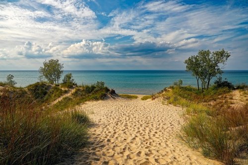 A sandy path leads to a calm lake, framed by green grass and trees under a blue sky with scattered clouds.
