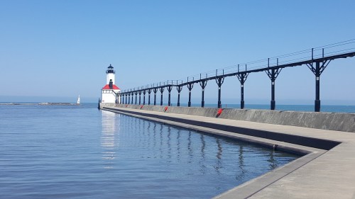 A lighthouse stands at the end of a long pier, with calm water and a clear blue sky in the background.