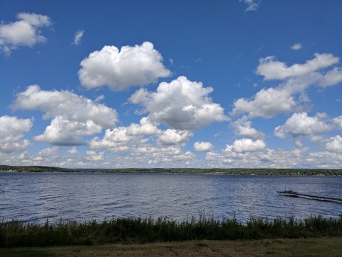 A serene lake view under a blue sky filled with fluffy white clouds, surrounded by green hills.