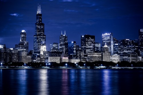 Chicago skyline at night, featuring illuminated skyscrapers reflecting on the water.