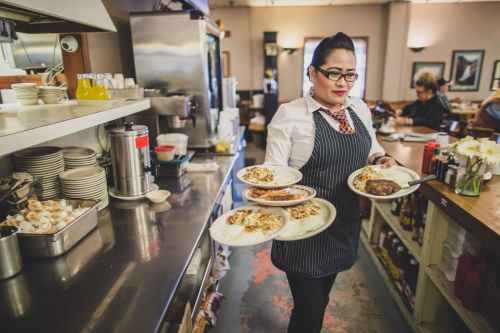 A waitress in a striped apron carries four plates of food in a bustling diner, with customers seated in the background.