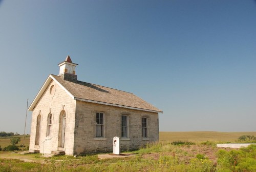 A small, historic stone schoolhouse stands alone in a grassy landscape under a clear blue sky.