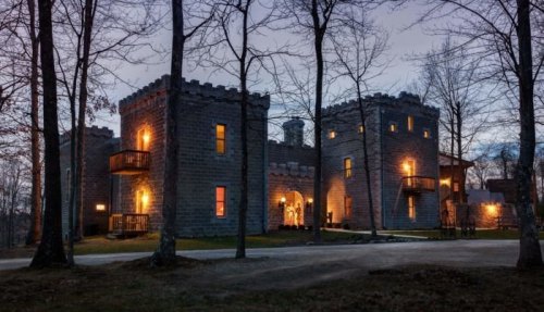 A stone castle with turrets, illuminated windows, surrounded by trees at dusk.