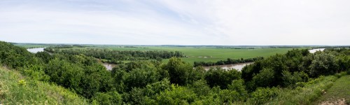 A panoramic view of lush green fields and winding rivers under a clear sky, surrounded by trees.