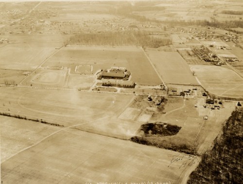 Aerial view of a rural area featuring fields, a large building, and scattered houses in the background.
