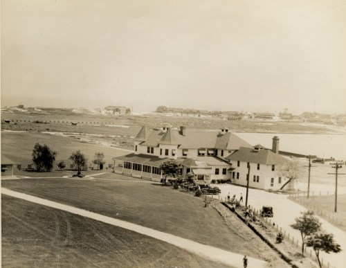 Historic black-and-white photo of a large house near a body of water, with a road and distant buildings in the background.