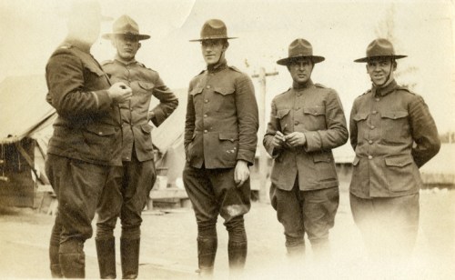 Five soldiers in military uniforms and hats stand together outdoors, with tents visible in the background.