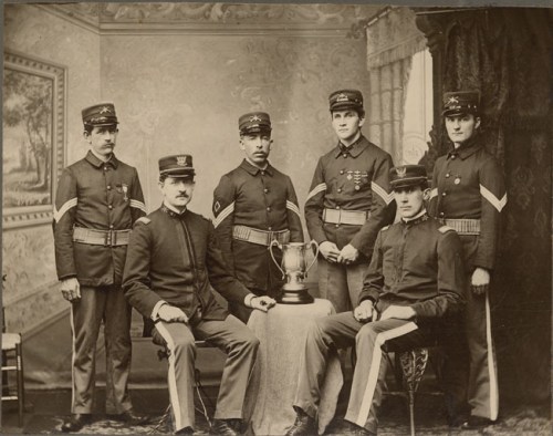 A historic black-and-white photo of six soldiers in uniform, posing around a trophy on a table.