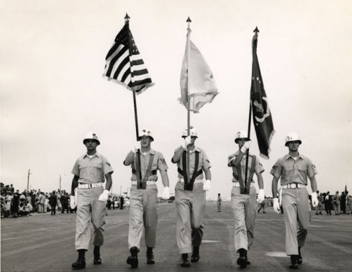 Five uniformed individuals march in formation, each holding a flag, with a crowd watching in the background.