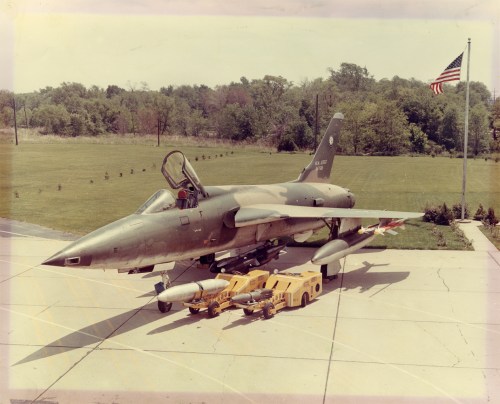 A military jet on a tarmac with missiles, set against a backdrop of trees and an American flag.