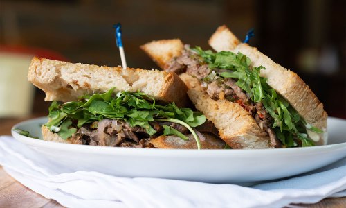 A close-up of two hearty sandwiches filled with greens and sliced meat, served on a white plate with toothpicks.