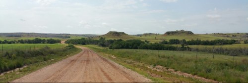 A dirt road winds through green fields, leading to distant hills under a partly cloudy sky.