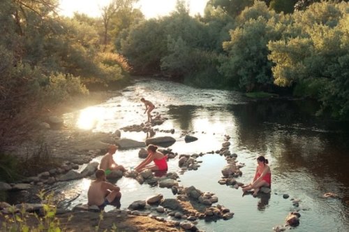 A group of people enjoying a sunny day by a river, sitting on rocks and playing in the water surrounded by greenery.