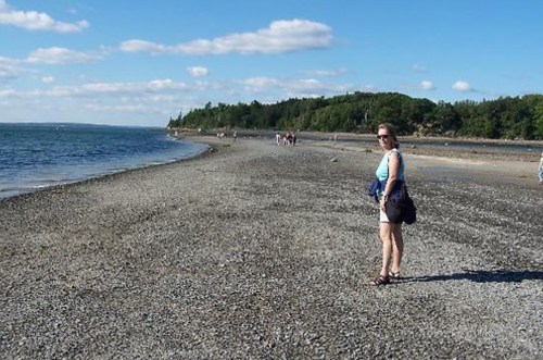 A woman stands on a rocky beach, looking back at the camera with a scenic shoreline and blue sky in the background.