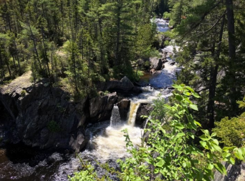 A scenic view of a waterfall cascading through rocky terrain, surrounded by lush green trees and a flowing river.