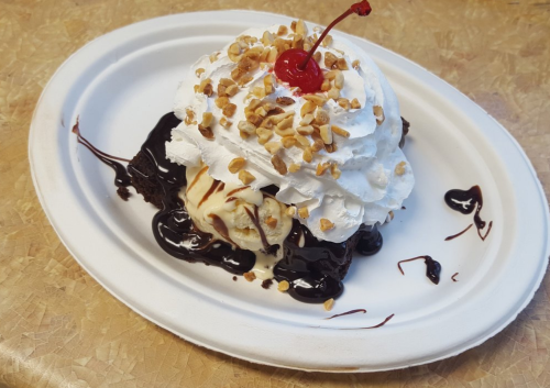 A dessert featuring vanilla ice cream topped with whipped cream, chopped nuts, chocolate sauce, and a cherry on a paper plate.