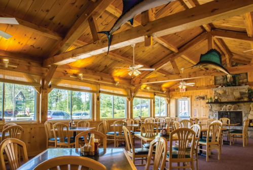 Cozy wooden restaurant interior with large windows, tables, and a stone fireplace, surrounded by nature.
