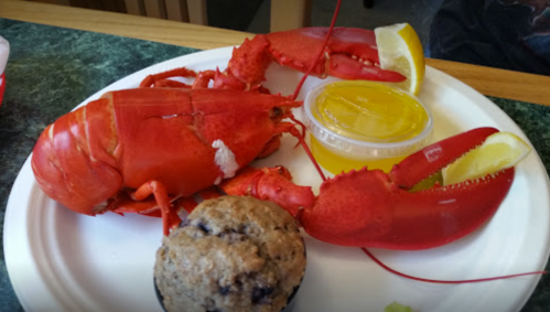 A plate with a cooked lobster, lemon wedges, melted butter, and a blueberry muffin.