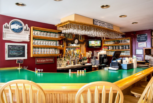 A cozy bar with a green countertop, shelves of drinks, and hanging glasses in a warm, inviting atmosphere.