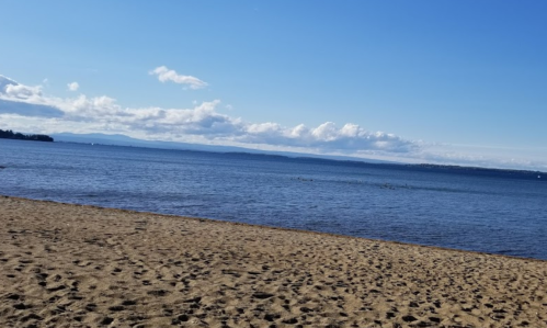 A sandy beach with calm water and distant mountains under a clear blue sky with scattered clouds.