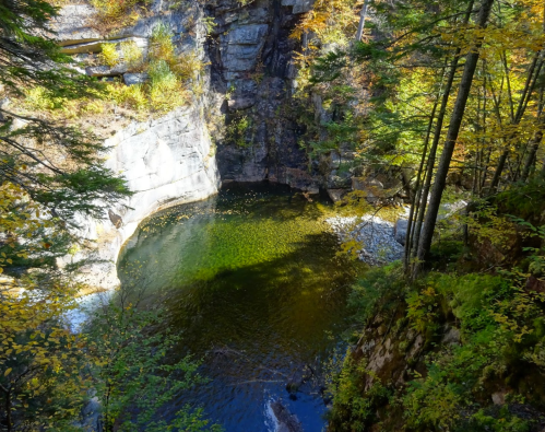A serene natural pool surrounded by rocky cliffs and autumn foliage, reflecting sunlight in a tranquil setting.