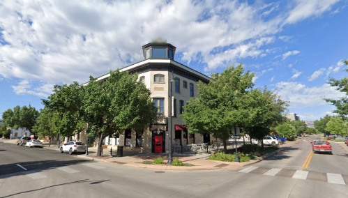 A corner building with a tower, surrounded by trees and parked cars, under a partly cloudy sky.