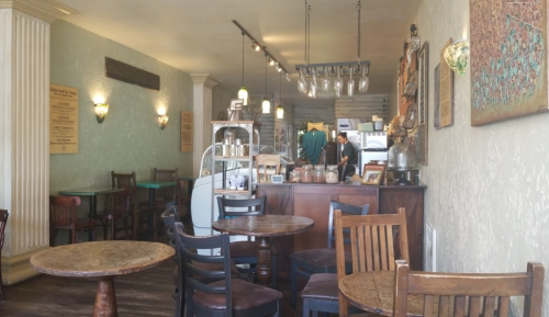 Cozy café interior with wooden tables, chairs, and a counter displaying baked goods and drinks. Warm lighting throughout.