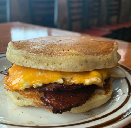 A close-up of a breakfast sandwich with bacon, cheese, and egg between two fluffy pancakes on a plate.