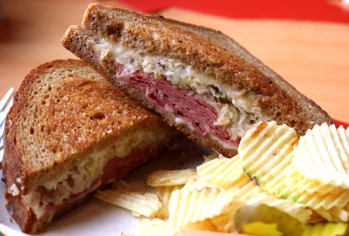 A close-up of a sliced sandwich with corned beef and sauerkraut on rye bread, served with a side of potato chips.
