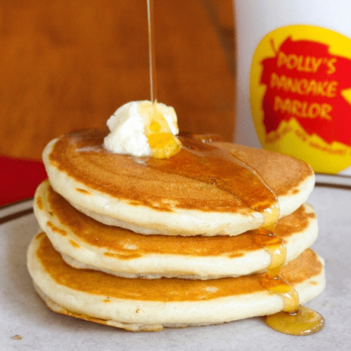 Three fluffy pancakes stacked on a plate, topped with butter and drizzled with syrup, next to a cup from a pancake parlor.