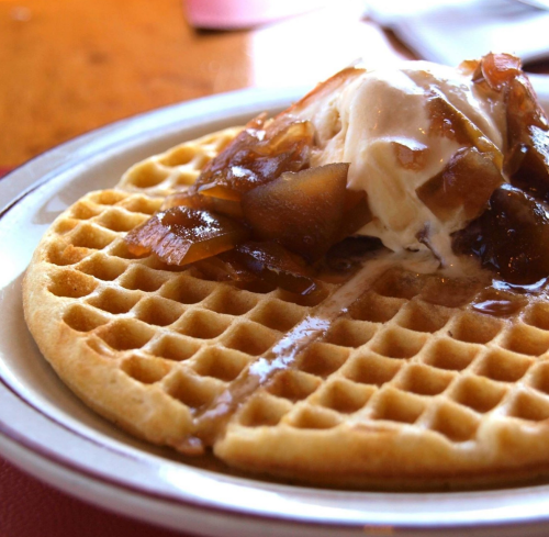 A golden waffle topped with ice cream and caramelized apples, served on a white plate.