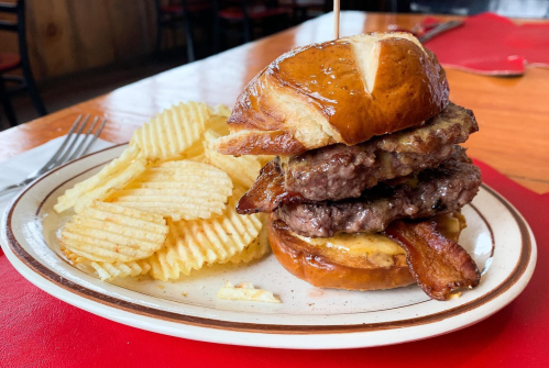 A stacked burger with two patties, bacon, and cheese on a pretzel bun, served with a side of ridged potato chips.