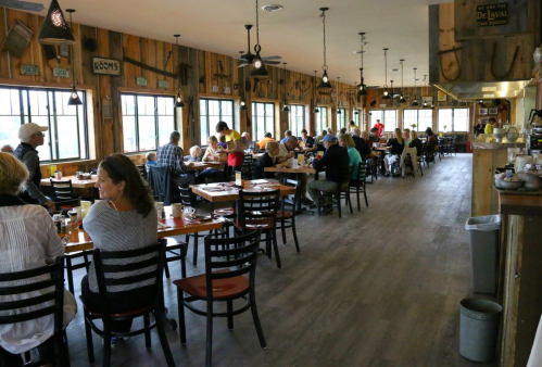 A bustling restaurant interior with wooden decor, filled with diners at tables and large windows letting in natural light.