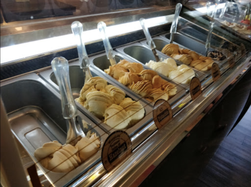 A display of various gelato flavors in metal containers, with scoops and labels visible.