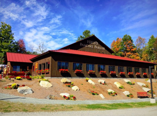 A rustic pancake parlor with a red roof, surrounded by colorful flowers and trees, under a blue sky.
