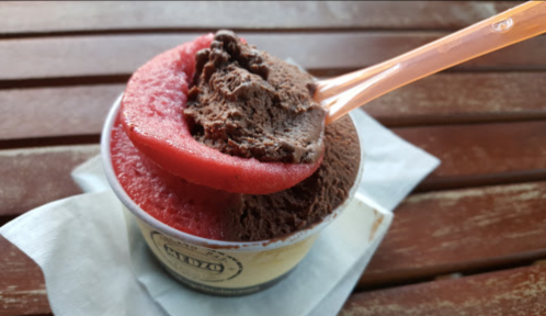 A cup of chocolate and strawberry gelato with a plastic spoon, resting on a napkin on a wooden table.