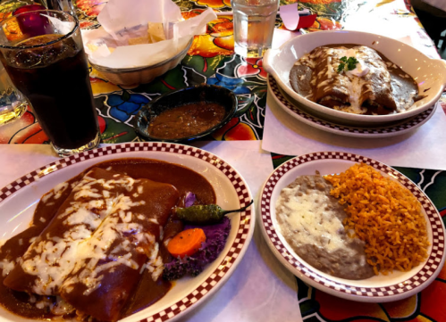 A colorful table setting with two plates of enchiladas, rice, refried beans, and a drink.