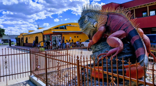 A large, colorful iguana sculpture sits behind a fence, with a yellow building and people in the background under a blue sky.