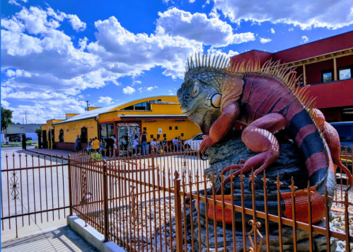 A large iguana sculpture sits in front of a colorful building, with people lined up nearby under a blue sky with clouds.