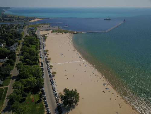 Aerial view of a sandy beach with people, cars lined along the road, and a calm blue lake in the background.