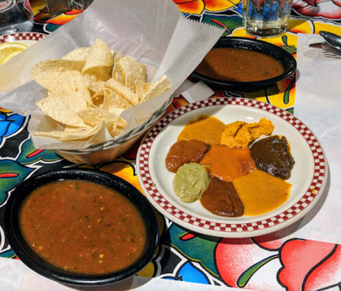 A basket of tortilla chips next to a plate with various colorful salsas and a bowl of salsa on a vibrant tablecloth.
