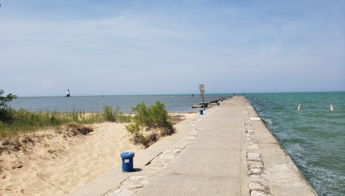 A concrete pier extends into a calm lake, with sandy beach on one side and a lighthouse in the distance.