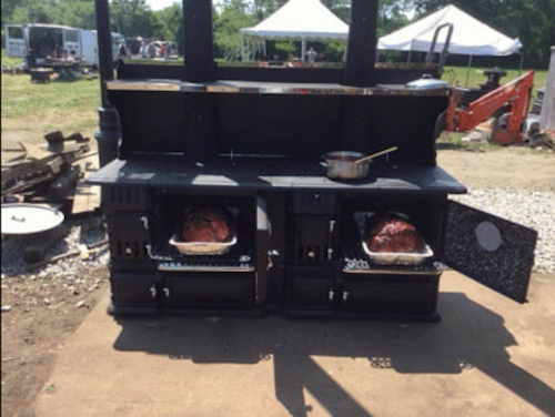 A large outdoor grill with two compartments, each holding a roasting meat, and a pot on top. Tents and equipment in the background.